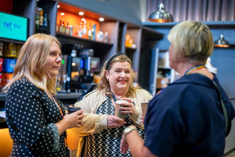 Three female DEI team members standing at chatting each holding disposable  coffee cups